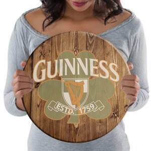 May include: A round wooden sign with a brown wood grain background. The sign features a green shamrock with a white harp inside. The text "GUINNESS" is in white letters above the shamrock and "ESTD. 1759" is in white letters below the shamrock.