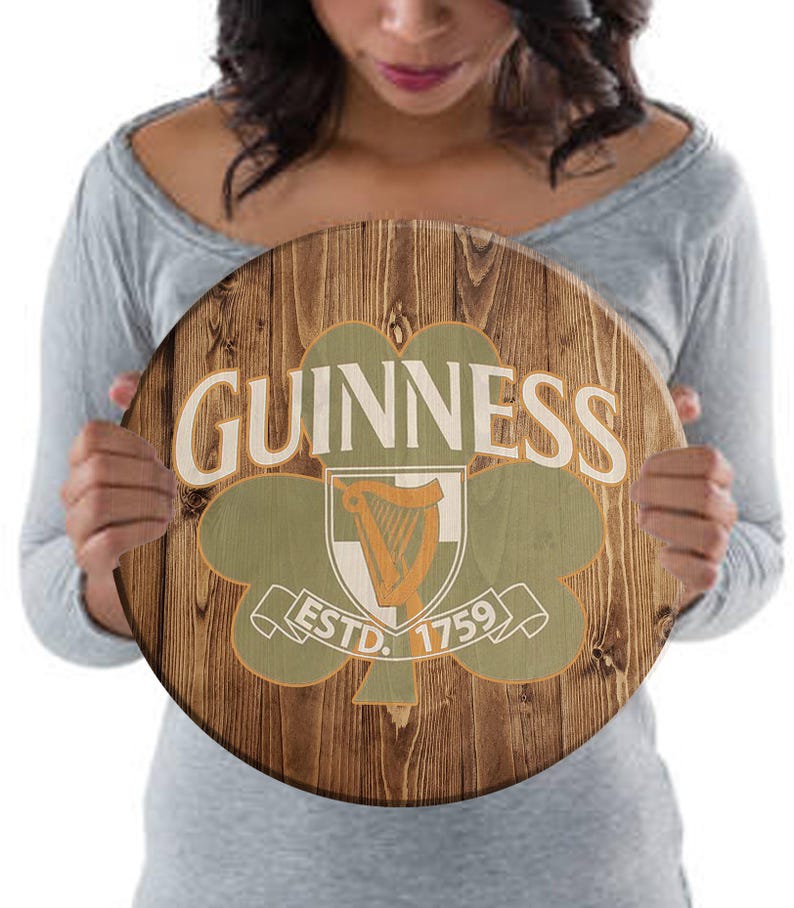 May include: A round wooden sign with a brown wood grain background. The sign features a green shamrock with a white harp inside. The text "GUINNESS" is in white letters above the shamrock and "ESTD. 1759" is in white letters below the shamrock.