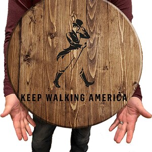 Puede incluir: Un letrero redondo de madera con una mancha marr&oacute;n. El letrero presenta una silueta negra de un hombre caminando con un bast&oacute;n y un sombrero de copa. El texto "KEEP WALKING AMERICA" est&aacute; impreso en negro debajo de la silueta.
