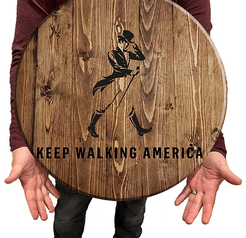 Puede incluir: Un letrero redondo de madera con una mancha marr&oacute;n. El letrero presenta una silueta negra de un hombre caminando con un bast&oacute;n y un sombrero de copa. El texto "KEEP WALKING AMERICA" est&aacute; impreso en negro debajo de la silueta.
