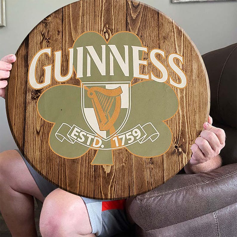 May include: A round wooden sign with a brown wood grain background. The sign features a green shamrock with a white harp inside a shield. The text "GUINNESS ESTD. 1759" is printed in white on the sign.
