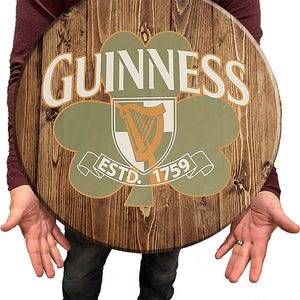 May include: A round wooden sign with a brown wood grain background. The sign features a green shamrock with a white harp inside. The text "GUINNESS ESTD. 1759" is printed in white on the sign.