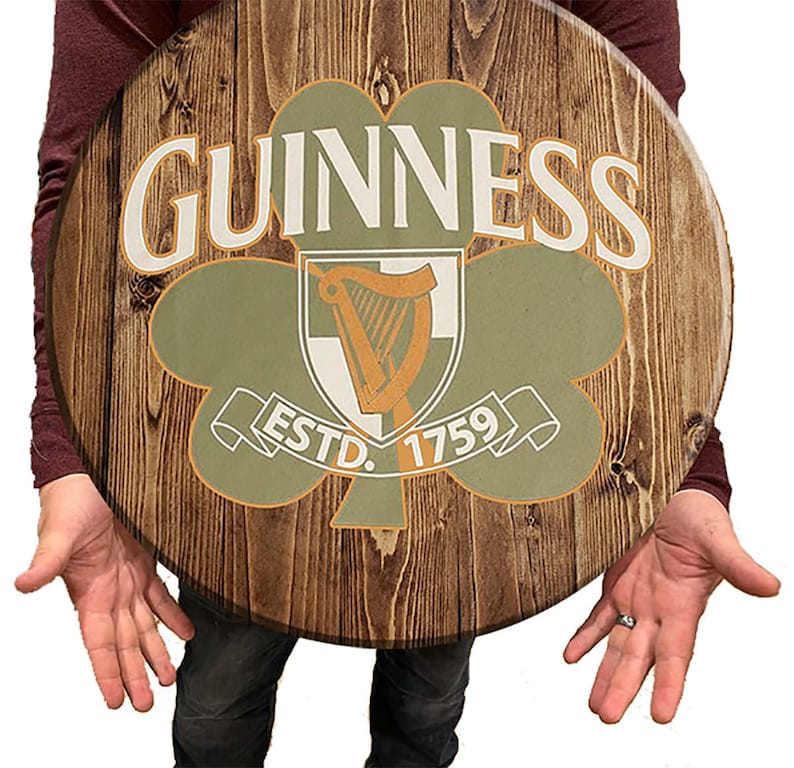 May include: A round wooden sign with a brown wood grain background. The sign features a green shamrock with a white harp inside. The text "GUINNESS ESTD. 1759" is printed in white on the sign.