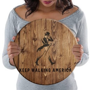 Puede incluir: Un letrero redondo de madera con un acabado de grano de madera marr&oacute;n. El letrero presenta una silueta negra de un hombre caminando con un bast&oacute;n y el texto "KEEP WALKING AMERICA".