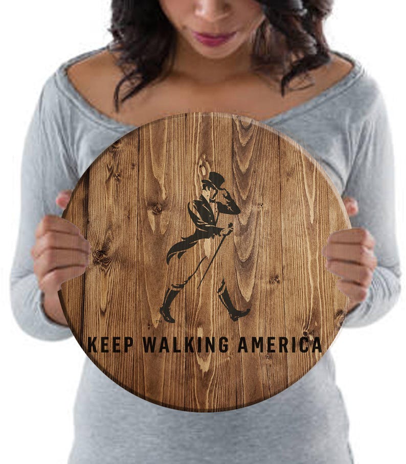 Puede incluir: Un letrero redondo de madera con un acabado de grano de madera marr&oacute;n. El letrero presenta una silueta negra de un hombre caminando con un bast&oacute;n y el texto "KEEP WALKING AMERICA".