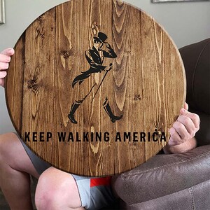 Puede incluir: Un letrero redondo de madera con una mancha marr&oacute;n. El letrero presenta una silueta negra de un hombre caminando con un bast&oacute;n y un sombrero de copa. El texto "KEEP WALKING AMERICA" est&aacute; impreso en negro debajo de la silueta.