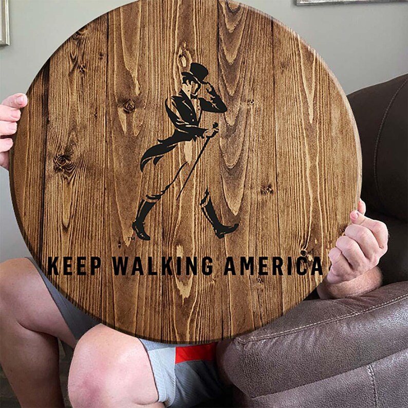 Puede incluir: Un letrero redondo de madera con una mancha marr&oacute;n. El letrero presenta una silueta negra de un hombre caminando con un bast&oacute;n y un sombrero de copa. El texto "KEEP WALKING AMERICA" est&aacute; impreso en negro debajo de la silueta.