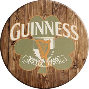 May include: A round wooden sign with a brown wood grain background. The sign features a green shamrock with a white harp inside. The words "GUINNESS ESTD. 1759" are printed on the sign.