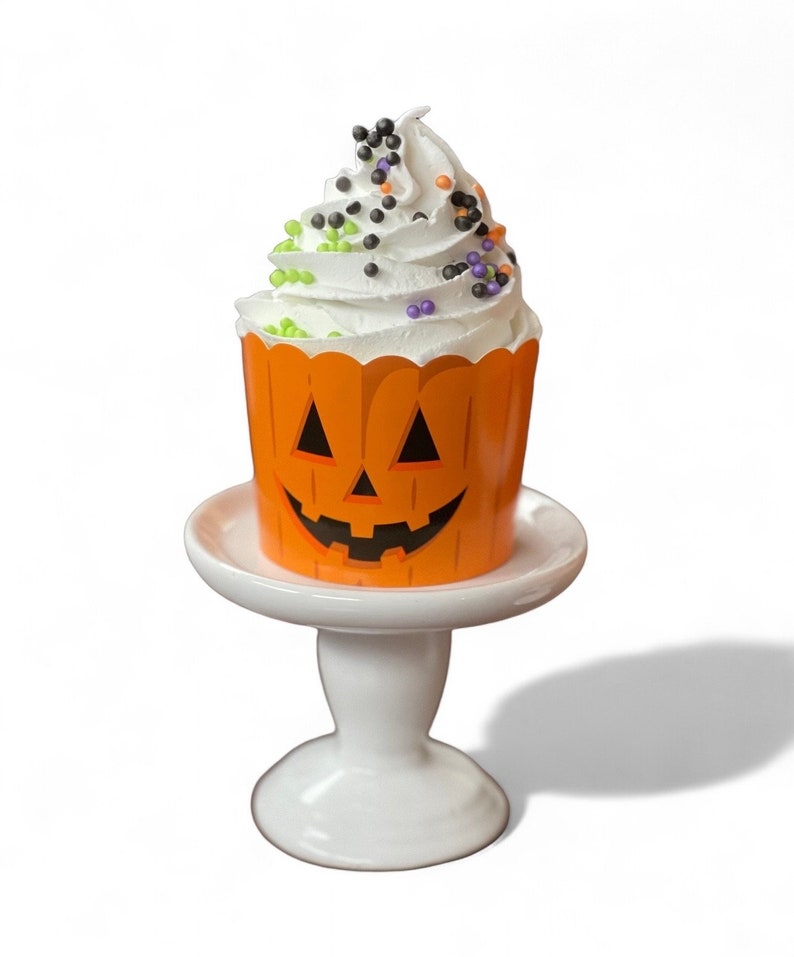 May include: A white cupcake with green, purple, and black sprinkles in a pumpkin-shaped orange cupcake liner on a white cake stand.