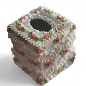 May include: White tissue box cover with pink and green floral accents. The cover is decorated with a ruffled design and pearl-like embellishments.