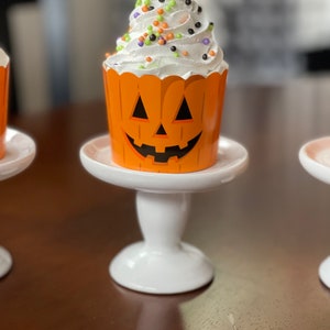 May include: A white cupcake with orange and black sprinkles, topped with white frosting, sitting on a white cake stand. The cupcake liner is decorated with a jack-o'-lantern face.