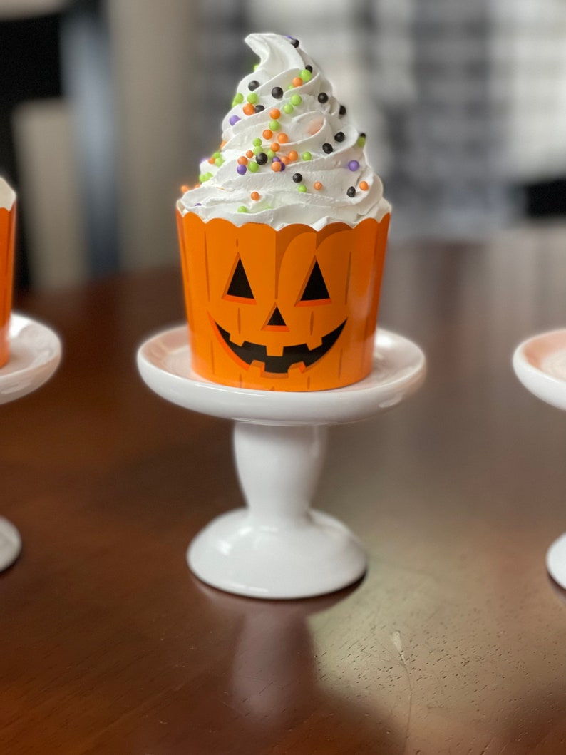 May include: A white cupcake with orange and black sprinkles, topped with white frosting, sitting on a white cake stand. The cupcake liner is decorated with a jack-o'-lantern face.