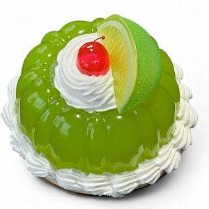 Op de afbeelding: Een groen gelei-dessert met een witte slagroomkern, gegarneerd met een rode kers en een limoenschijfje. Het dessert is omgeven door een ring van witte slagroom. Het dessert staat op een ronde, lichtbruine basis.