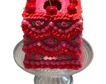 Può includere: Un copri scatola di fazzoletti rosa e rosso a forma di torta con ciliegie in cima. La torta è decorata con glassa rosa e bianca e accenti perlati.