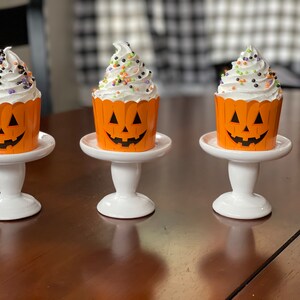 May include: Three white cupcakes with orange pumpkin-shaped wrappers and black and green sprinkles are displayed on white cake stands. The cupcakes are on a brown wooden table.
