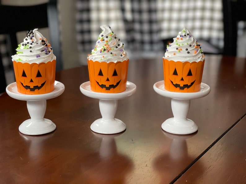 May include: Three white cupcakes with orange pumpkin-shaped wrappers and black and green sprinkles are displayed on white cake stands. The cupcakes are on a brown wooden table.