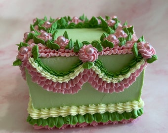 Può includere: Una torta quadrata verde decorata con glassa rosa e verde, con un bordo a balze e dettagli di perle.