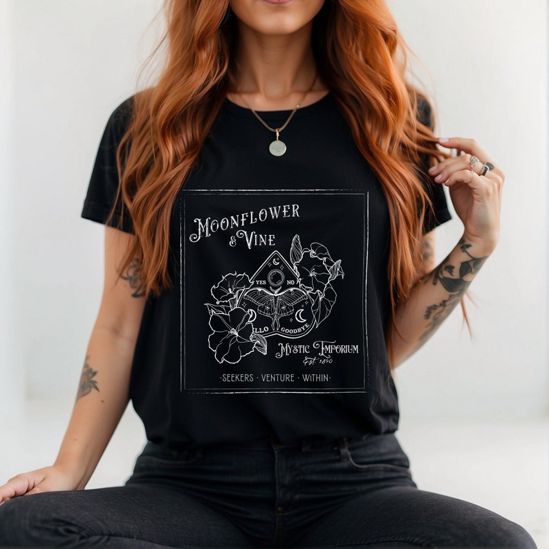 Pode incluir: Camiseta preta com um design gr&aacute;fico branco que apresenta uma flor de lua e uma videira, um tabuleiro Ouija e o texto "Moonflower & Vine Mystic Emporium est. 2020 Seekers Venture Within".