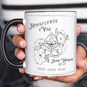 May include: White ceramic mug with a black handle. The mug features a black and white illustration of a moonflower, vine, and a Ouija board with a moth. The text reads "Moonflower & Vine Mystic Emporium Est. 1890 Seekers Venture Within."