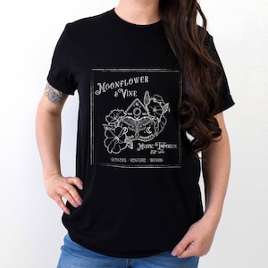 Pode incluir: Camiseta preta com um design gr&aacute;fico branco que apresenta uma flor de lua e uma videira, um tabuleiro Ouija e o texto "Moonflower & Vine Mystic Emporium Est. 2010 Seekers Venture Within".