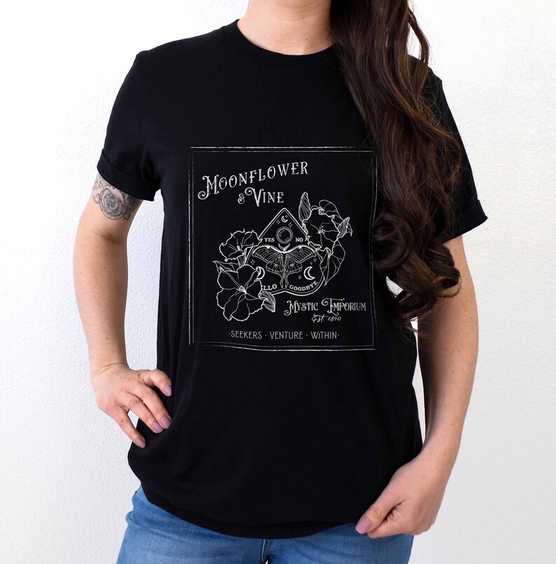 Pode incluir: Camiseta preta com um design gr&aacute;fico branco que apresenta uma flor de lua e uma videira, um tabuleiro Ouija e o texto "Moonflower & Vine Mystic Emporium Est. 2010 Seekers Venture Within".