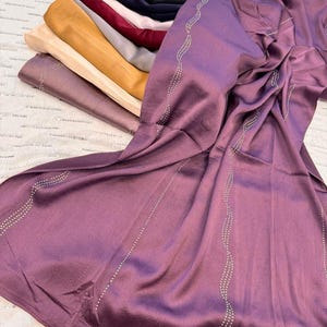 May include: A collection of satin scarves in various colors, including purple, gold, and navy. The primary scarf is a deep purple with a decorative, wavy line of small, white embellishments. The scarves are neatly stacked, showcasing their smooth texture.