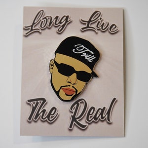 May include: A black and white enamel pin featuring a cartoon portrait of a man wearing a black baseball cap with the word "Tru" written in white on the front of the cap. The man is wearing black sunglasses and has a brown beard and mustache. The pin has the words "Long Live The Real" written in a stylized font around the portrait.