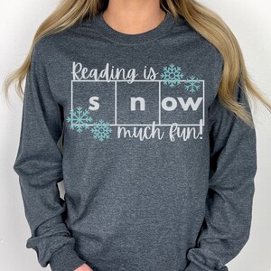 May include: A dark heather grey long-sleeve shirt with the text "Reading is snow much fun!" in white lettering. The text is in a box with snowflakes on either side.