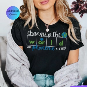 May include: Black t-shirt with the text "changing the world one phoneme at a time" in white letters. The text is in a box with the letters "w", "o", "r", "l", and "d" in individual boxes. A blue heart is in the "o" box and a green globe is above the text.