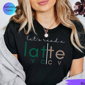 May include: Black t-shirt with the phrase "let's read a latte" in white, green, and brown lettering. The shirt has a crew neck and short sleeves. Additional colors are available. The word "Black" is written in a purple box.