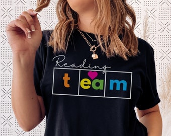 Peut inclure: T-shirt noir avec l'inscription "Reading Team" en écriture blanche. En dessous, le mot "team" est dans un encadré avec les lettres orange, vertes et bleues, et un cœur rose. Le t-shirt est de style décontracté.