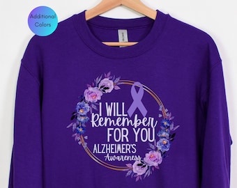 Może przedstawiać: Fioletowa bluza z napisem "I WILL Remember FOR YOU Alzheimer's Awareness" wewnątrz kwiatowego wieńca i fioletowej wstążki. Bluza wisi na drewnianym wieszaku.