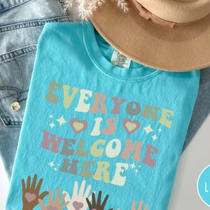 May include: A turquoise blue t-shirt with the text "Everyone is Welcome Here" in a retro font with rainbow colours. The text is surrounded by small stars and hearts. The bottom of the shirt features a design of multiple hands of different skin tones holding hearts.