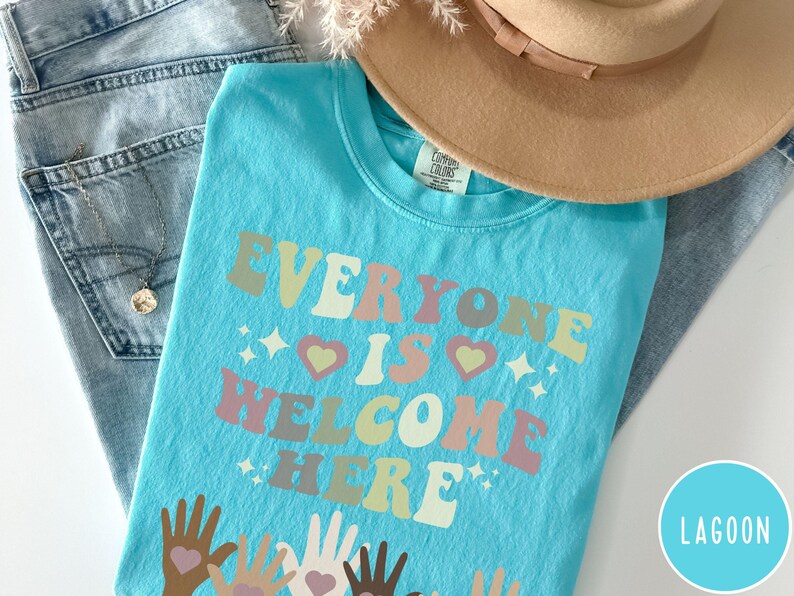 May include: A turquoise blue t-shirt with the text "Everyone is Welcome Here" in a retro font with rainbow colours. The text is surrounded by small stars and hearts. The bottom of the shirt features a design of multiple hands of different skin tones holding hearts.