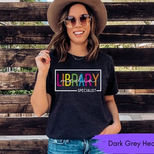 May include: Dark grey heather t-shirt with the text "LIBRARY SPECIALIST" in a colorful rainbow font.