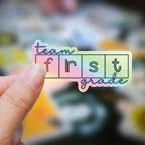 May include: A holographic sticker with the words "team first grade" in a playful font. The sticker has a pastel rainbow gradient background and is held by a hand. The design is suitable for teachers or students.
