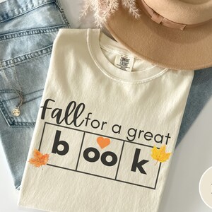 Puede incluir: Camiseta de color marfil con el texto "fall for a great book" en letras negras. La palabra "book" est&aacute; en un cuadro con un coraz&oacute;n en lugar de la "o". Hay peque&ntilde;as hojas naranjas y amarillas a cada lado del cuadro.