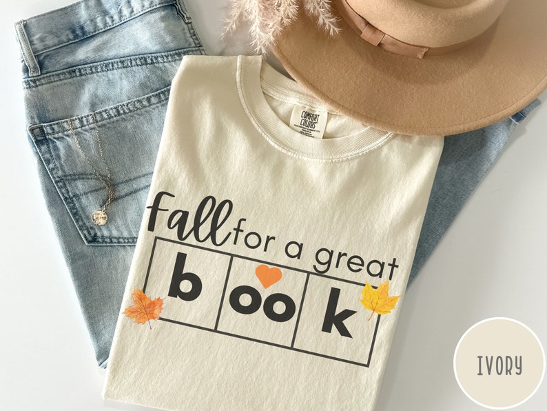 Puede incluir: Camiseta de color marfil con el texto "fall for a great book" en letras negras. La palabra "book" est&aacute; en un cuadro con un coraz&oacute;n en lugar de la "o". Hay peque&ntilde;as hojas naranjas y amarillas a cada lado del cuadro.