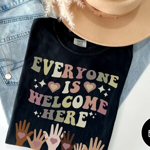 May include: Black t-shirt with the text "Everyone is Welcome Here" in rainbow colours. The text is in a retro font style. There are multiple hands with hearts at the bottom of the image.