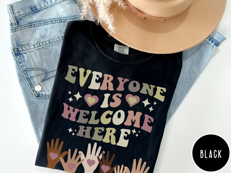 May include: Black t-shirt with the text "Everyone is Welcome Here" in rainbow colours. The text is in a retro font style. There are multiple hands with hearts at the bottom of the image.