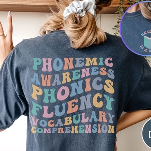 May include: A dark blue t-shirt with the words "PHONEMIC AWARENESS PHONICS FLUENCY VOCABULARY COMPREHENSION" in rainbow colors on the back. The front of the shirt has a small graphic of a book with the text "READING IS A RIGHT".