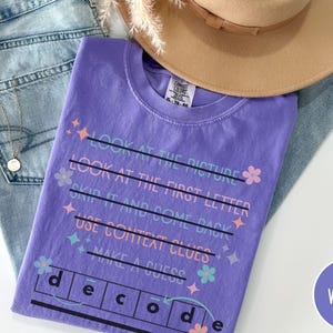 May include: A violet t-shirt with the word "decode" and instructions printed on it. The shirt is laid on top of a pair of light blue jeans, with a tan hat and a gold necklace. The shirt has the word "VIOLET" in a circle.