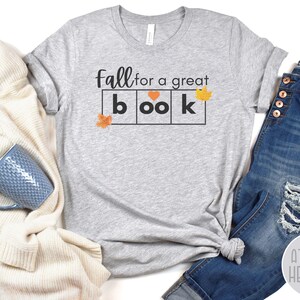 May include: Grey t-shirt with the text "Fall for a great book" in black lettering. The word "book" is in a block letter style with a heart in the "o". There are small orange leaf graphics on the shirt.