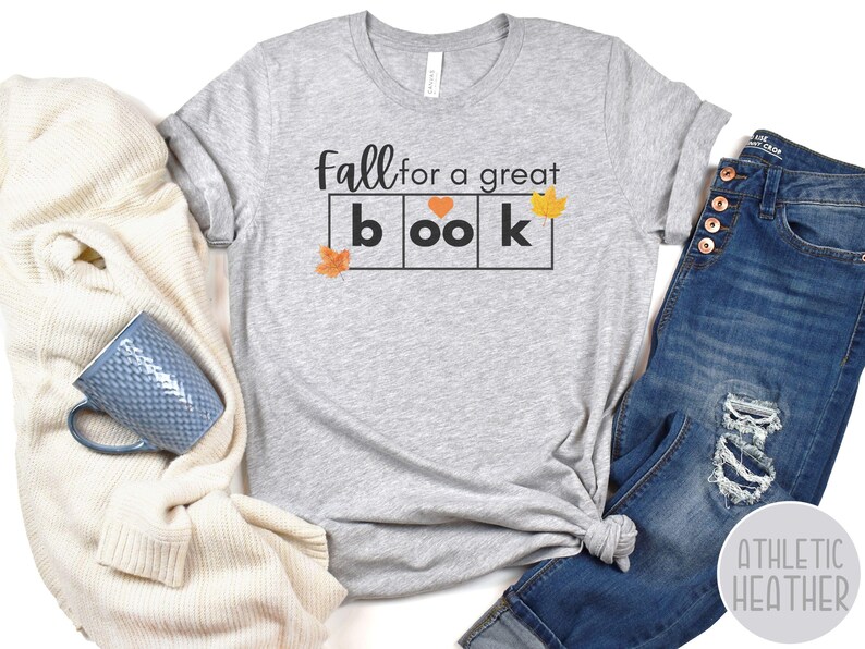 May include: Grey t-shirt with the text "Fall for a great book" in black lettering. The word "book" is in a block letter style with a heart in the "o". There are small orange leaf graphics on the shirt.