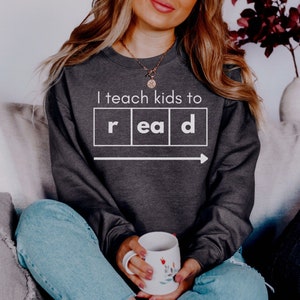 May include: A dark gray sweatshirt with the text "I teach kids to read" and a graphic of the word "read" in boxes with an arrow pointing to the right.