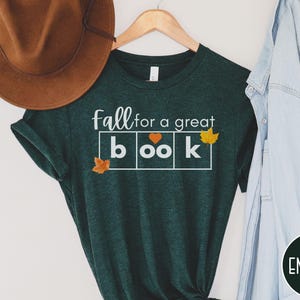 May include: A dark green t-shirt with the text "fall for a great book" in white letters. The word "book" is in a box with a heart in the "o". There are small orange leaf graphics on the shirt.