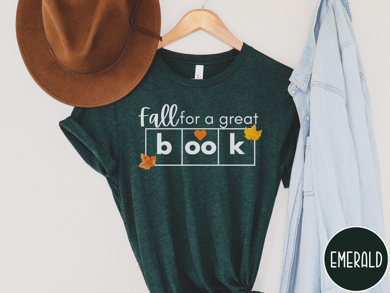 May include: A dark green t-shirt with the text "fall for a great book" in white letters. The word "book" is in a box with a heart in the "o". There are small orange leaf graphics on the shirt.