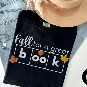 Puede incluir: Camiseta negra con el texto "Fall for a great book" en letras blancas. La palabra "book" est&aacute; en un cuadro con un coraz&oacute;n en lugar de la "o". Hay tres hojas de oto&ntilde;o en la camiseta.