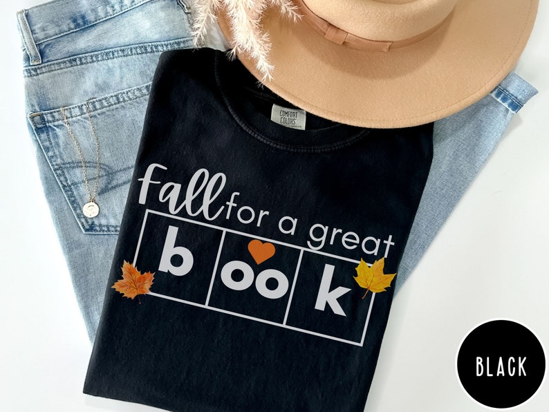 Puede incluir: Camiseta negra con el texto "Fall for a great book" en letras blancas. La palabra "book" est&aacute; en un cuadro con un coraz&oacute;n en lugar de la "o". Hay tres hojas de oto&ntilde;o en la camiseta.