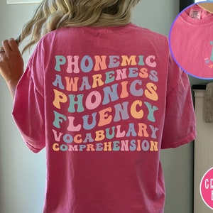May include: Pink t-shirt with the words "phonemic awareness, phonics, fluency, vocabulary, comprehension" in rainbow colors on the back. The front of the shirt has a small book icon with the text "Reading is a right".